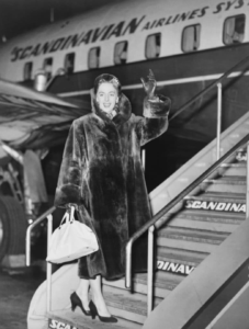 Photograph of Jorgensen in a large fur coat smiling and waving from the staircase to an airplane