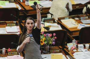 Montana Rep. Zooey Zephyr holding a microphone high above her head with her other hand in a fist against her chest