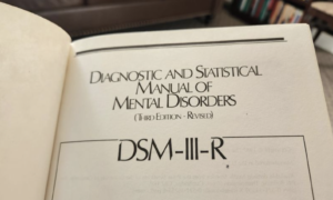 Close-up photograph of an open copy of the Diagnostic and Statistical Manual of Mental Disorders (DSM-III-R). The manual reflects the history of psychiatric classification and debates over medical recognition, diagnosis, and the pathologization of transgender identities.