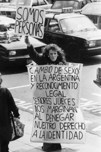 Black and white photograph of activist Karina Urbina holding signs demanding legal recognition of gender identity. One sign reads "Somos personas" ("We are people"), while another translates to “Gender transition and legal recognition in Argentina. Judges marginalize us by denying our right to identity.”