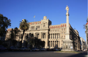 A wide daytime view of the Buenos Aires City Legislature shows the grand neoclassical government building with rows of tall columns, an Argentine flag flying above the roof, and the monument to General Juan Lavalle standing in the foreground. This building served as the site of major demonstrations in 2004 protesting reforms that expanded police authority over public space and disproportionately targeted marginalized communities.