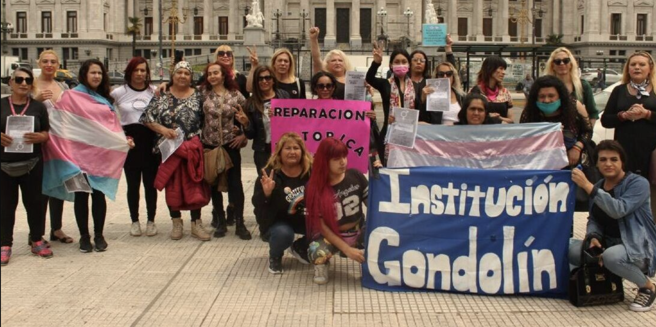 Color photograph of travesti and trans activists gathered in front of Argentina's National Congress holding trans pride flags and banners reading "Hotel Gondolín" and "Historic Reparations."