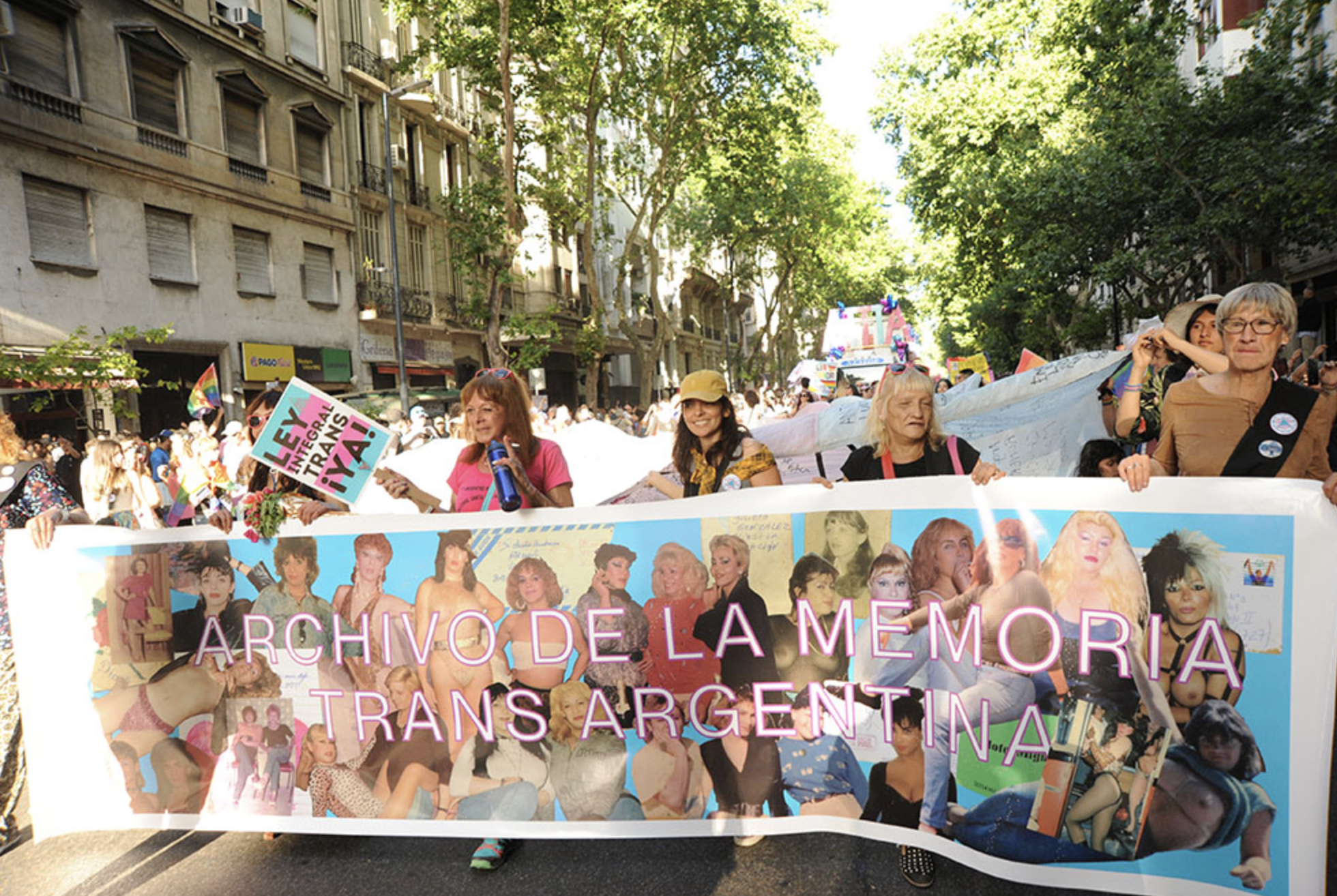 A large group of marchers proceeds down a tree-lined street in Buenos Aires carrying a wide banner for the Archivo de la Memoria Trans Argentina (Argentine Trans Memory Archive). The banner is covered with archival photographs of travestis and trans women spanning several decades. Participants of different ages walk together behind the banner, while one person holds a sign reading "LEY INTEGRAL TRANS ¡YA!" ("Comprehensive Trans Law Now!")