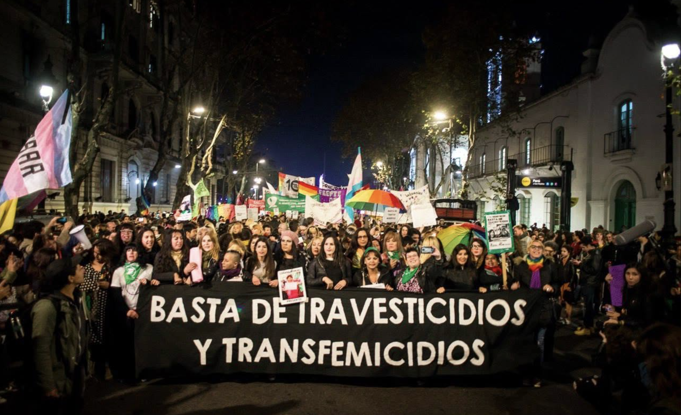Color photograph of hundreds of people marching at night through the streets of Buenos Aires behind a large banner reading "Basta de Travesticidios y Transfemicidios" ("Stop Travesticides and Transfemicides"). Photo by Nuria Boda (2018) via Emergentes
