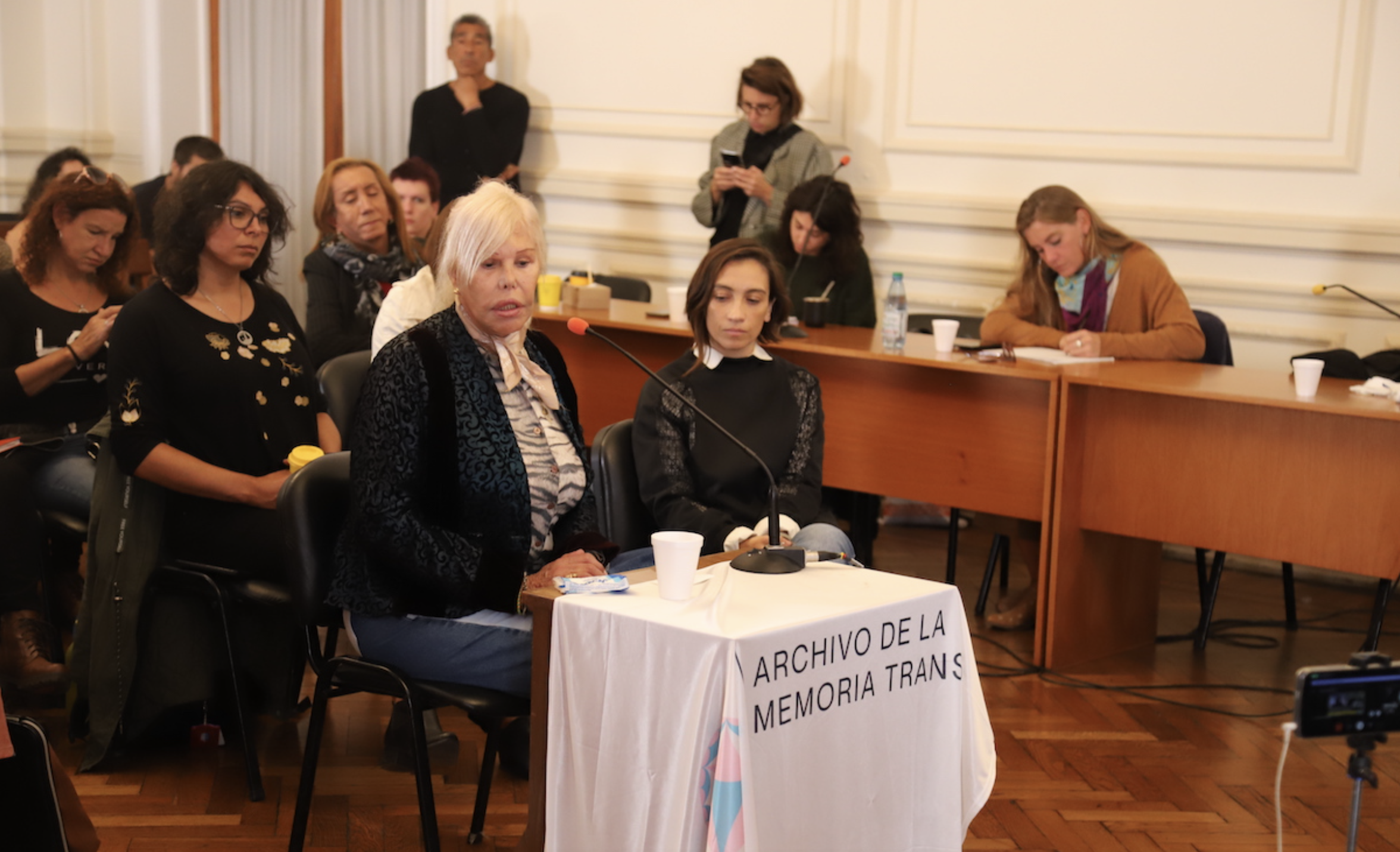 Analía Velázquez testifies in the Brigadas trial in 2024 seated at a table draped with a banner of the Trans Memory Archive which collaborated with the investigation by providing data on trans and travesti survivors of State Terrorism. Via Agencia Presentes