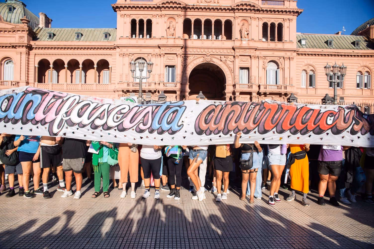 Color photograph of demonstrators gathered in front of the Casa Rosada in Buenos Aires holding a large banner reading "Antifascista, Antirracista" ("Anti-Fascist, Anti-Racist").